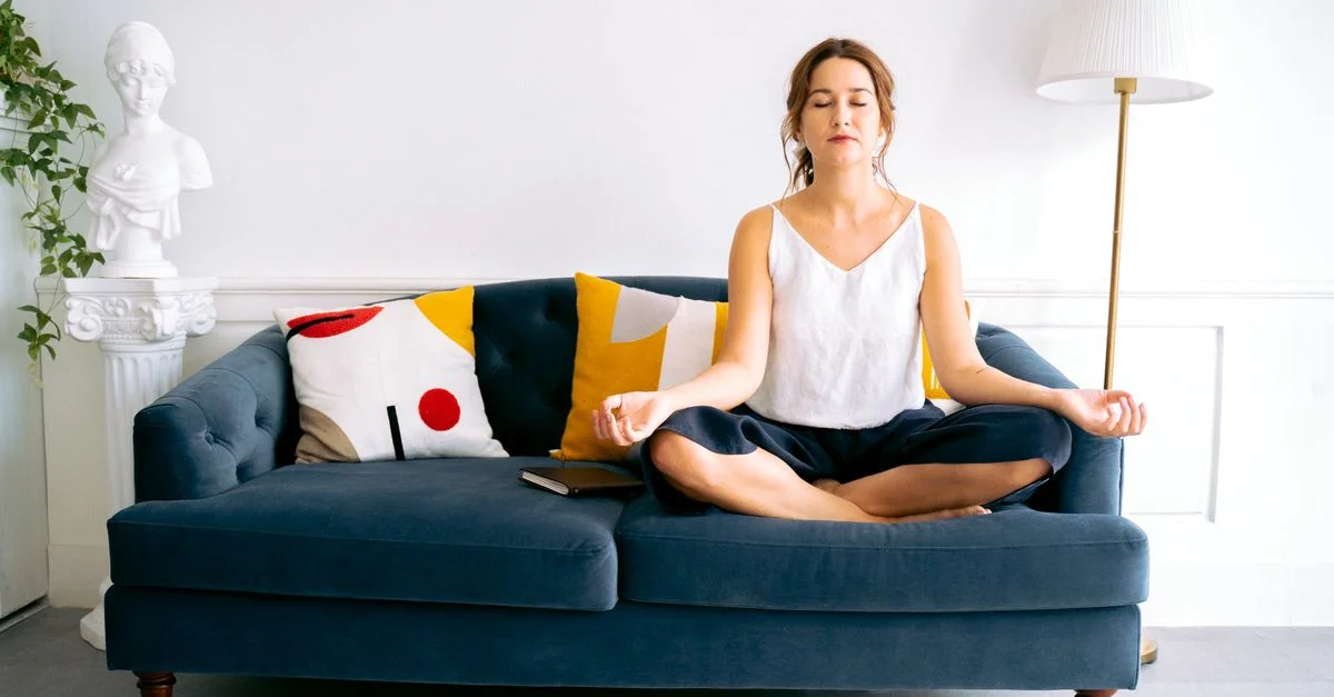 Femme en pose du lotus méditant sur un canapé dans un salon moderne lumineux.