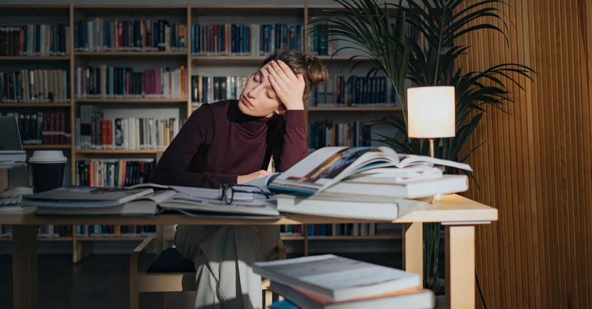 Une femme fatiguée entourée de livres, étudiant dans une bibliothèque, se sentant surmenée et stressée.