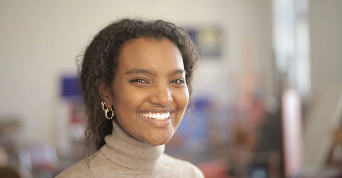Une femme joyeuse avec des cheveux bouclés souriant à l'intérieur, portant un pull à col roulé.