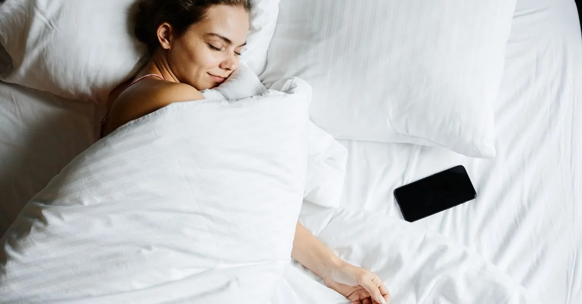 Une jeune femme dort confortablement dans son lit avec un smartphone à ses côtés, enveloppée dans des draps blancs.