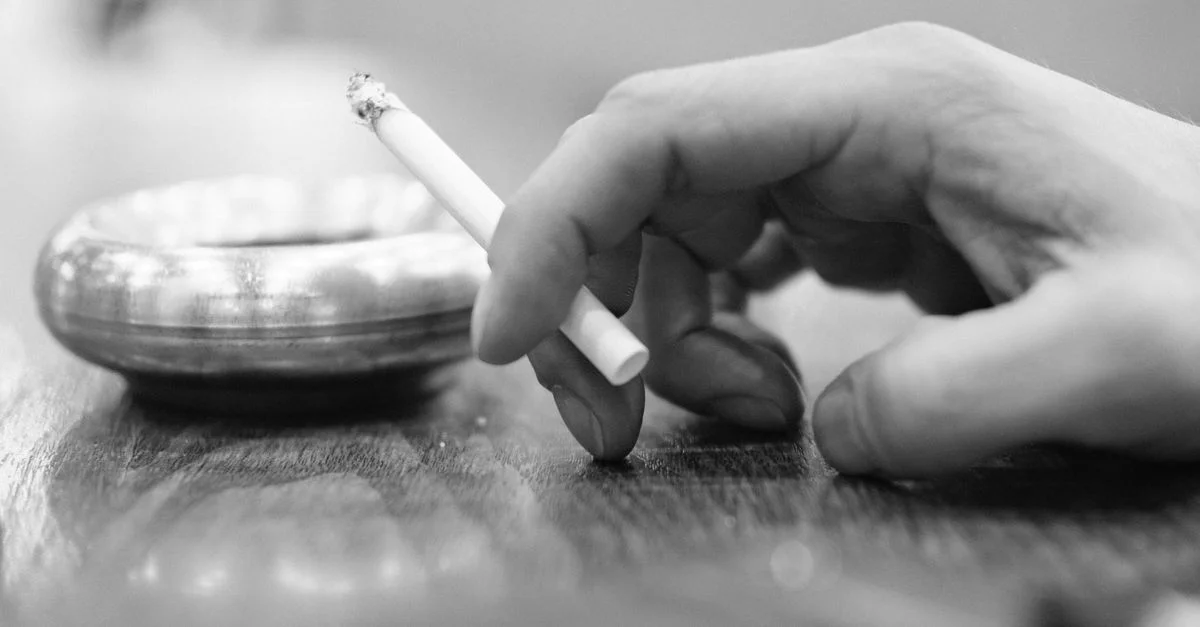 Photo monochrome montrant une main tenant une cigarette à côté d'un cendrier.