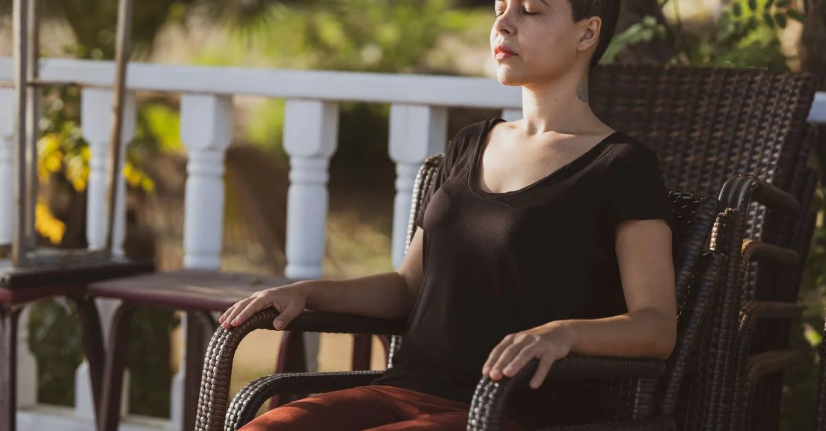 Une femme en méditation, les yeux fermés, assise sur une chaise à l'extérieur sous la lumière du soleil.